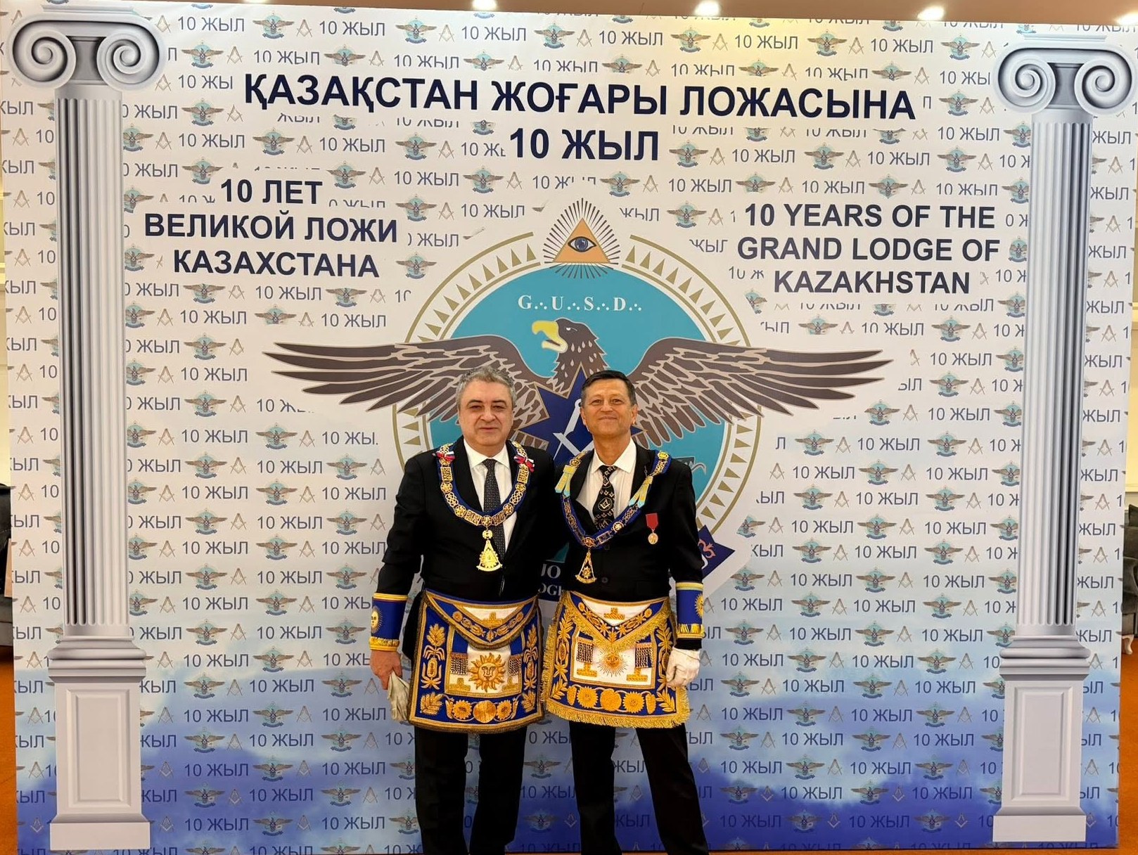 Two Freemasons in regalia before the banner of the tenth anniversary of the Grand Lodge of Kazakhstan